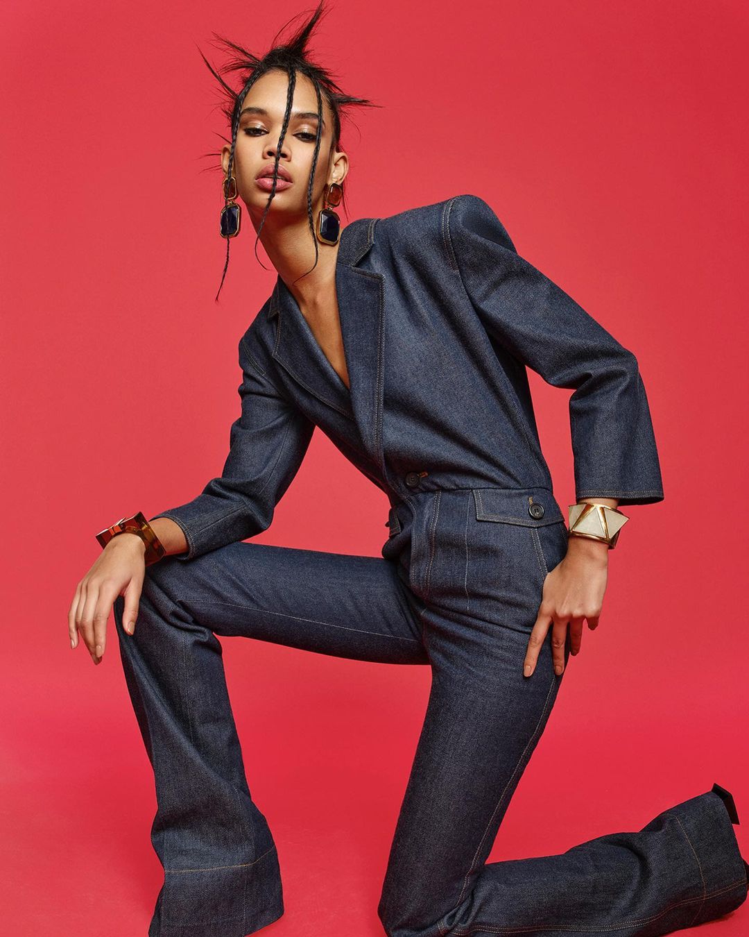 A fashionable adult female poses kneeling on a red background while wearing a denim jumpsuit with statement earrings and a large geometric cuff bracelet.