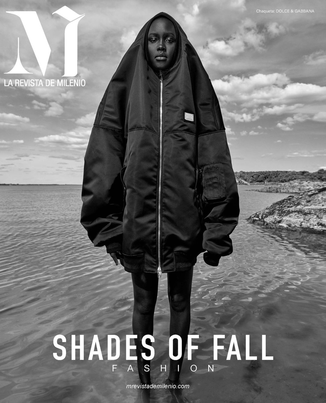 Fashion editorial of a tall Black woman wearing an oversized black hooded coat standing in shallow water against a cloudy sky and distant coastline.