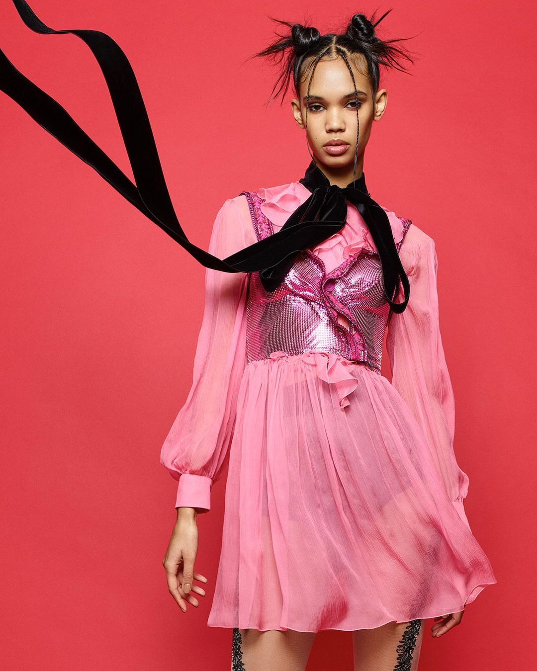 Portrait of a young adult woman with space buns, wearing a shiny pink dress with a velvet ribbon tie, against a coral background.