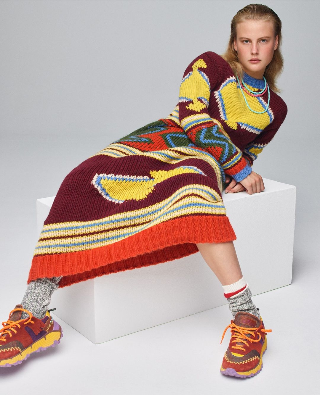 A woman poses in a vibrant, patterned knit dress and athletic shoes, seated on a white box against a neutral background in a studio setting.