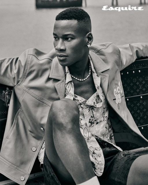 Portrait of a stylish, dark-skinned adult male in an unbuttoned jacket and floral shirt, sitting outdoors. A modern and fashionable look.