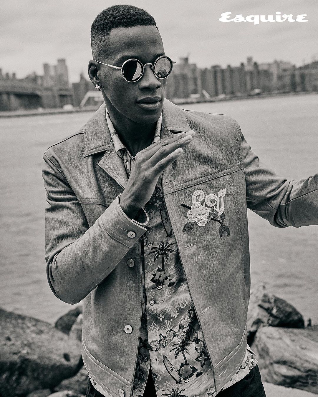 Monochrome portrait of a Black adult male wearing sunglasses and a light jacket, with a cityscape and body of water in the backdrop.
