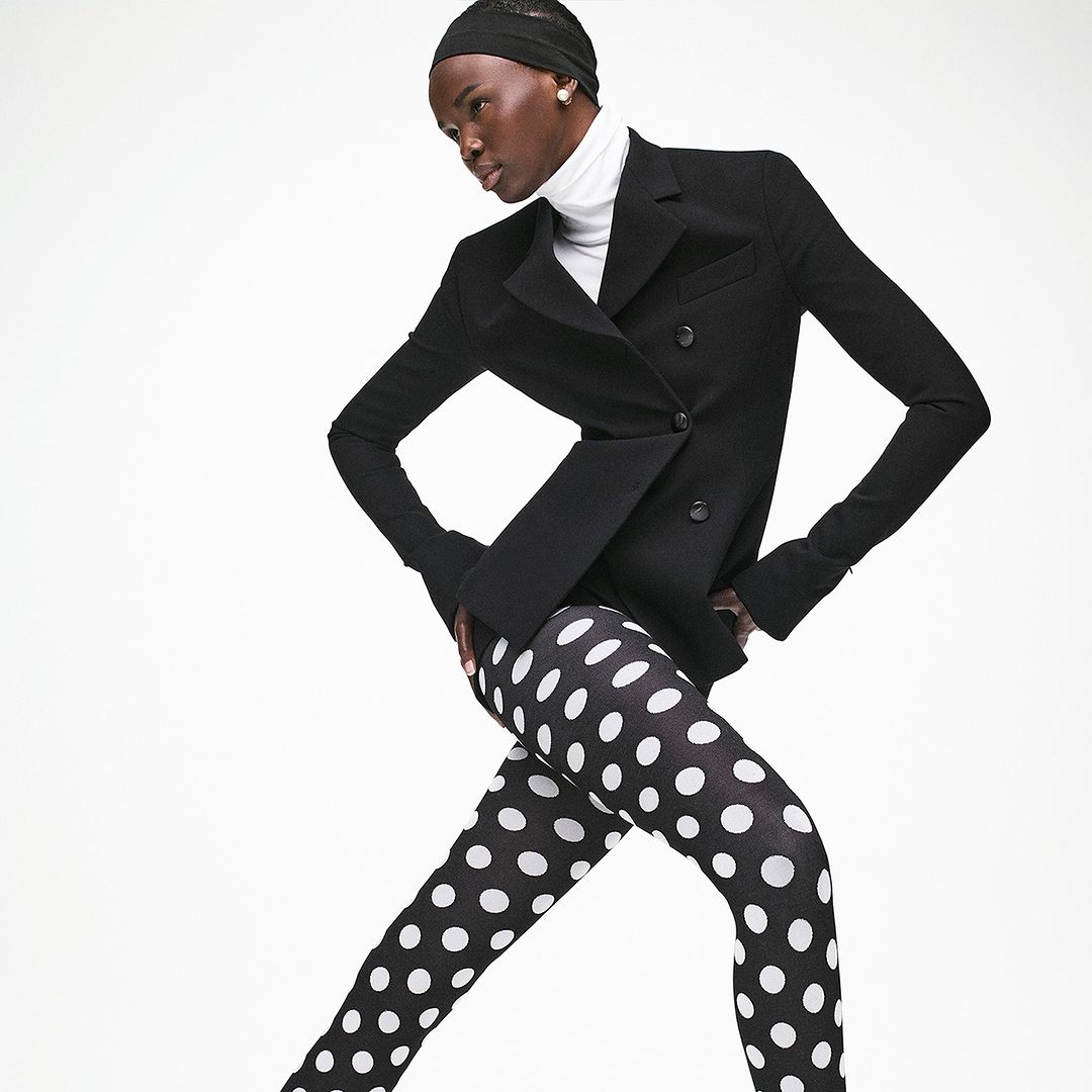 A striking fashion shot of a Black female model in polka dot tights, a white turtleneck, and a black blazer, against a bright white backdrop.