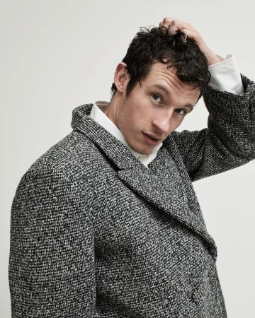 Man wearing a textured grey tweed coat and white collared shirt running his hand through his short dark hair against a plain grey background