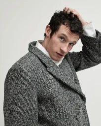 Man wearing a textured grey tweed coat and white collared shirt running his hand through his short dark hair against a plain grey background