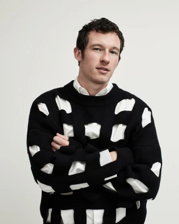 Man with curly brown hair wearing a black cutout sweater over a white shirt against a white background.