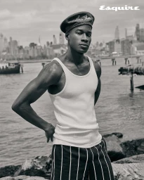 Black man wearing a white tank top, striped trousers, and beret against an urban waterfront background.