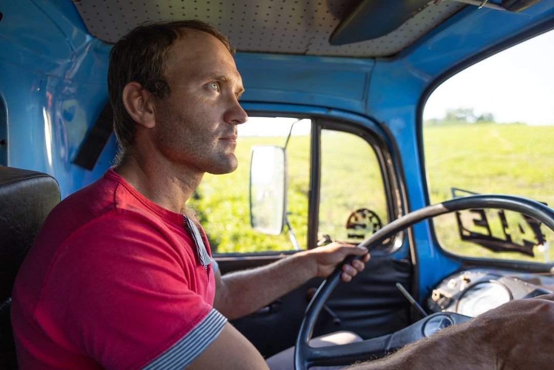 Adult man driving a vintage blue truck through the countryside, wearing a red shirt, looking off to the side with a concentrated gaze.