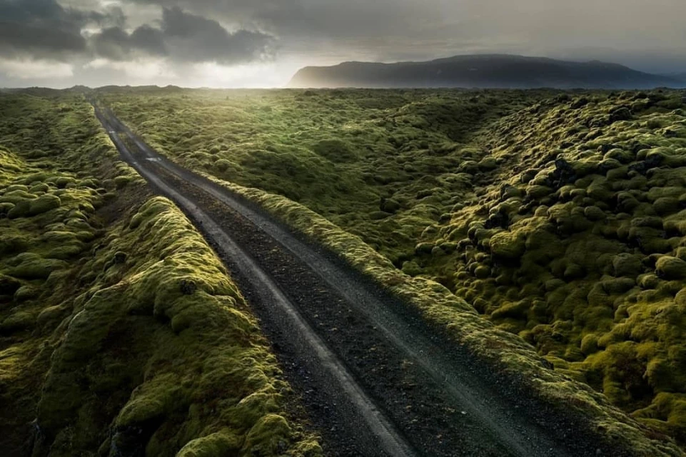 Overhead aerial view of a rugged black gravel road cutting through undulating green moss hills, under an overcast sky in Iceland.