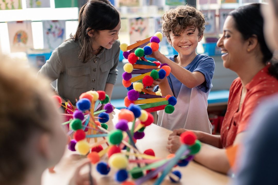 Smiling children and teacher at table in classroom working on a DNA molecule model made of colored pom poms and craft sticks.