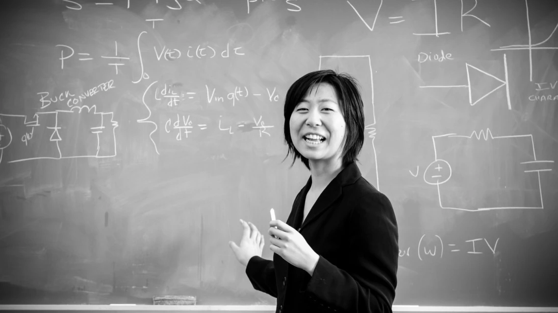 Asian woman standing near a blackboard covered in mathematical equations and diagrams in a classroom, wearing a dark blazer, smiling.