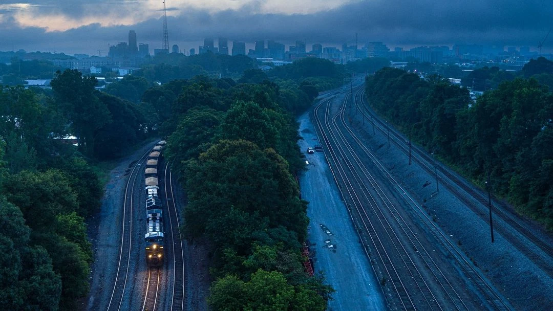 Aerial view of a freight train on a curved track surrounded by green trees approaching a distant city skyline at dusk or dawn.
