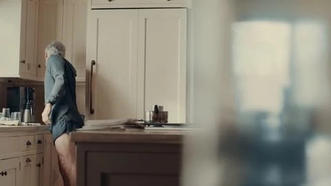 Adult male standing in a kitchen with light wood cabinets, wearing a blue shirt and shorts. A coffee maker sits on the counter.