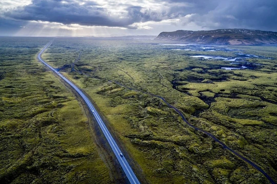 Aerial view of a straight road cutting through a vast green mossy landscape under a cloudy sky, leading to distant hills.