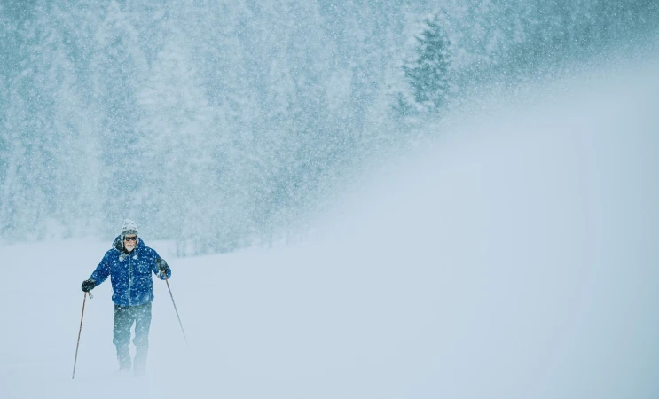 Elderly adult cross-country skiing on a snowy trail through a forest during a blizzard, wearing a blue jacket and a beanie.