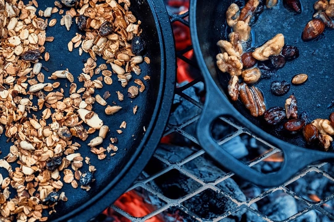 Granola and mixed nuts are cooked in cast iron pans over an open campfire, providing a rustic and delicious outdoor meal.