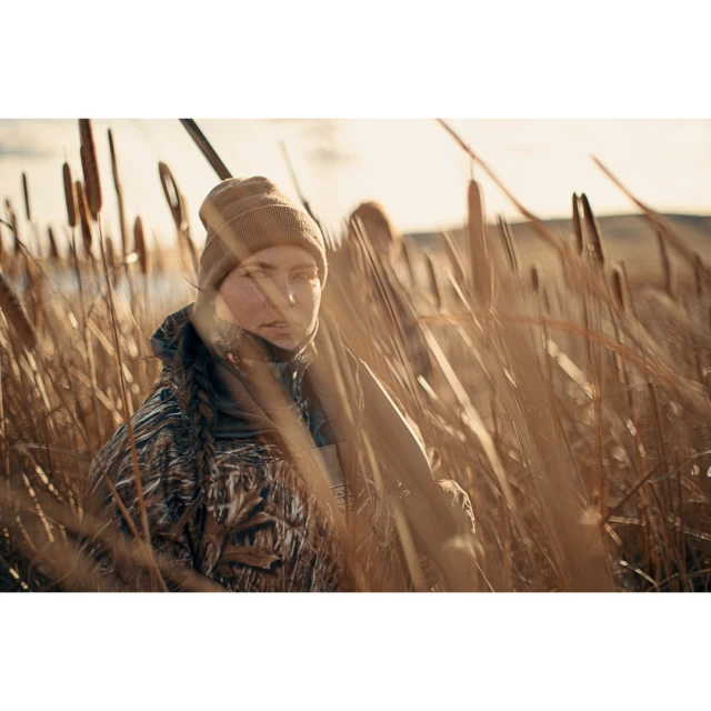 Woman in camouflage and knit cap with a hunting rifle poses in a field of cattails under a bright sky at eye level.