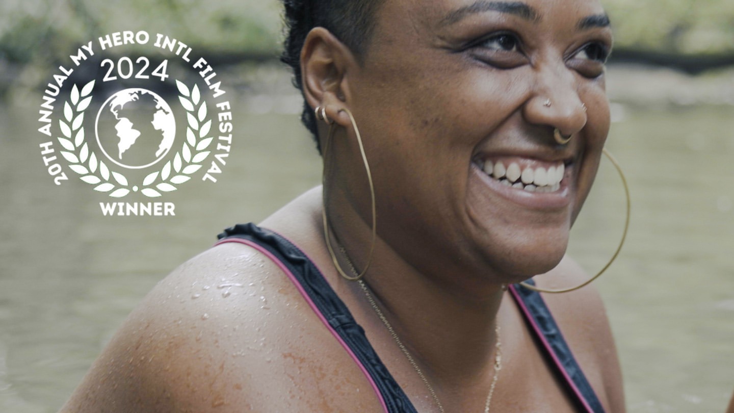 Smiling woman with hoop earrings near water, wearing a black bathing suit with pink trim. The 20th Annual My Hero Intl Film Festival logo.
