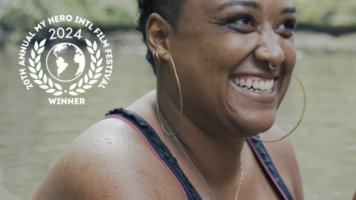 Smiling woman with hoop earrings near water, wearing a black bathing suit with pink trim. The 20th Annual My Hero Intl Film Festival logo.