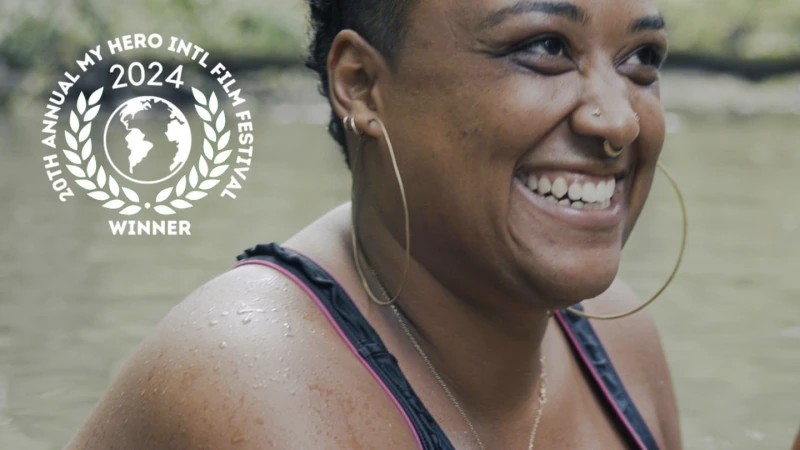 Smiling woman with hoop earrings near water, wearing a black bathing suit with pink trim. The 20th Annual My Hero Intl Film Festival logo.