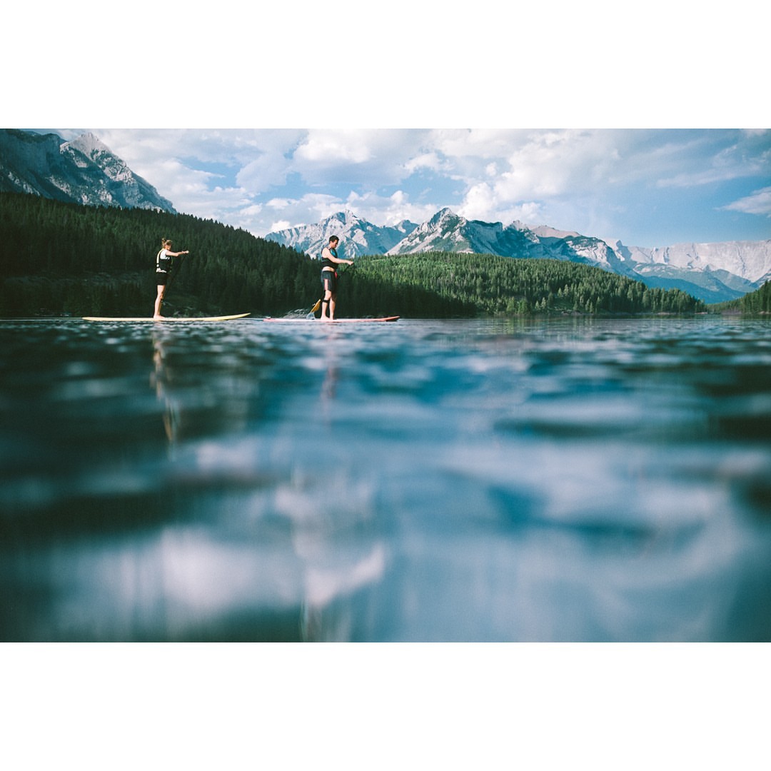 Two people paddleboarding on a lake surrounded by a forest and snow-capped mountains under a partly cloudy sky on a sunny day.