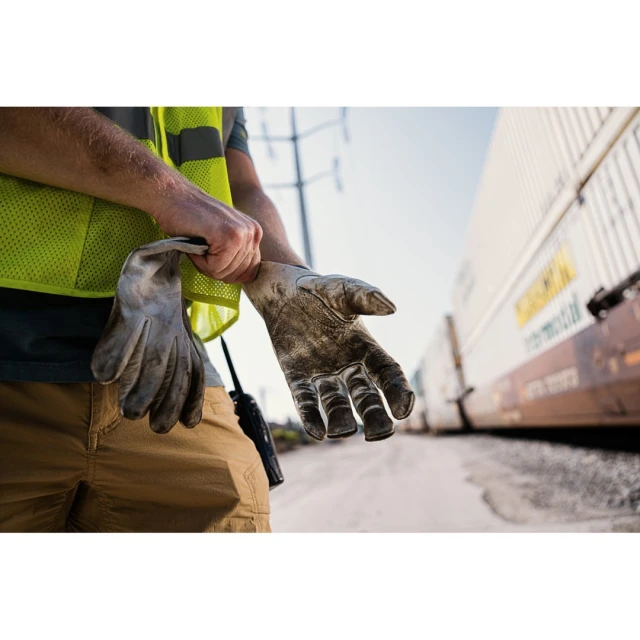 Construction worker wearing safety vest and gloves near train cars with tan pants, shot outside during daylight, with a medium shot.