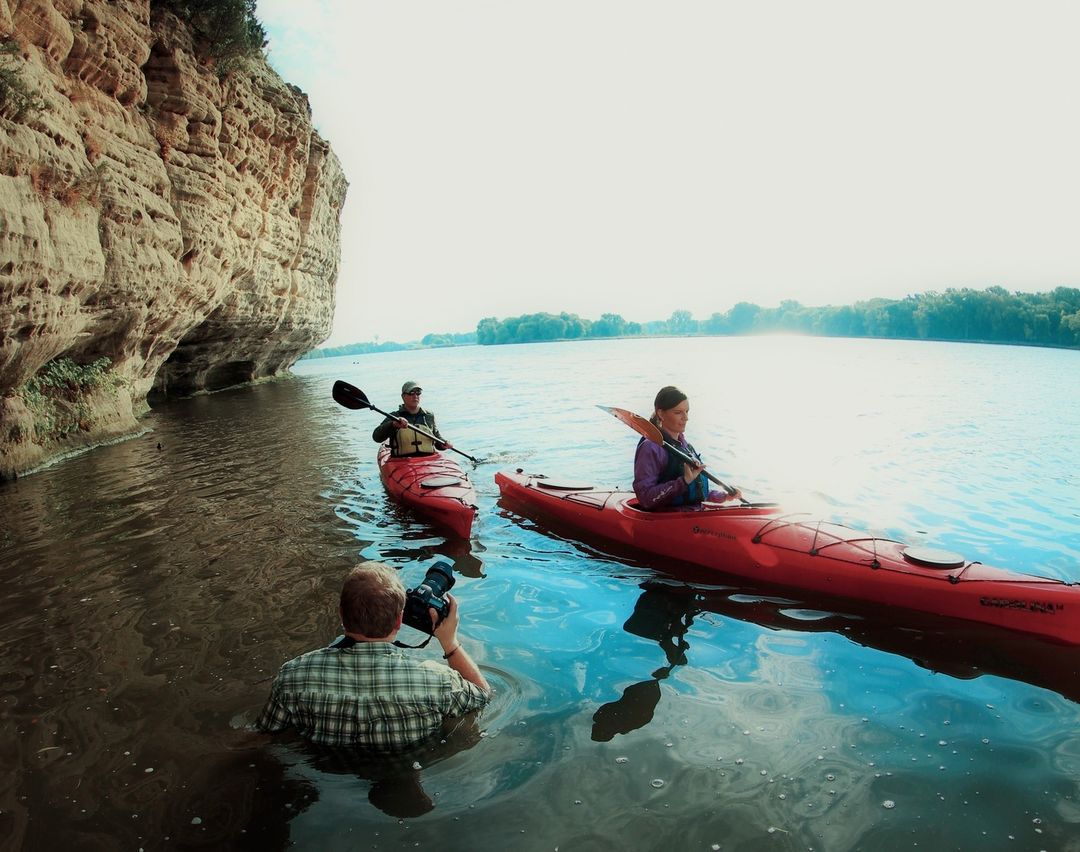 Two women paddle red kayaks on a river, as a man in water takes photos near a rocky cliff, under bright skies. Water reflects the light.