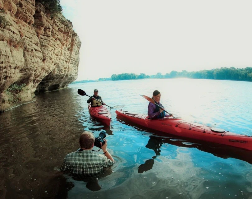 Two women paddle red kayaks on a river, as a man in water takes photos near a rocky cliff, under bright skies. Water reflects the light.