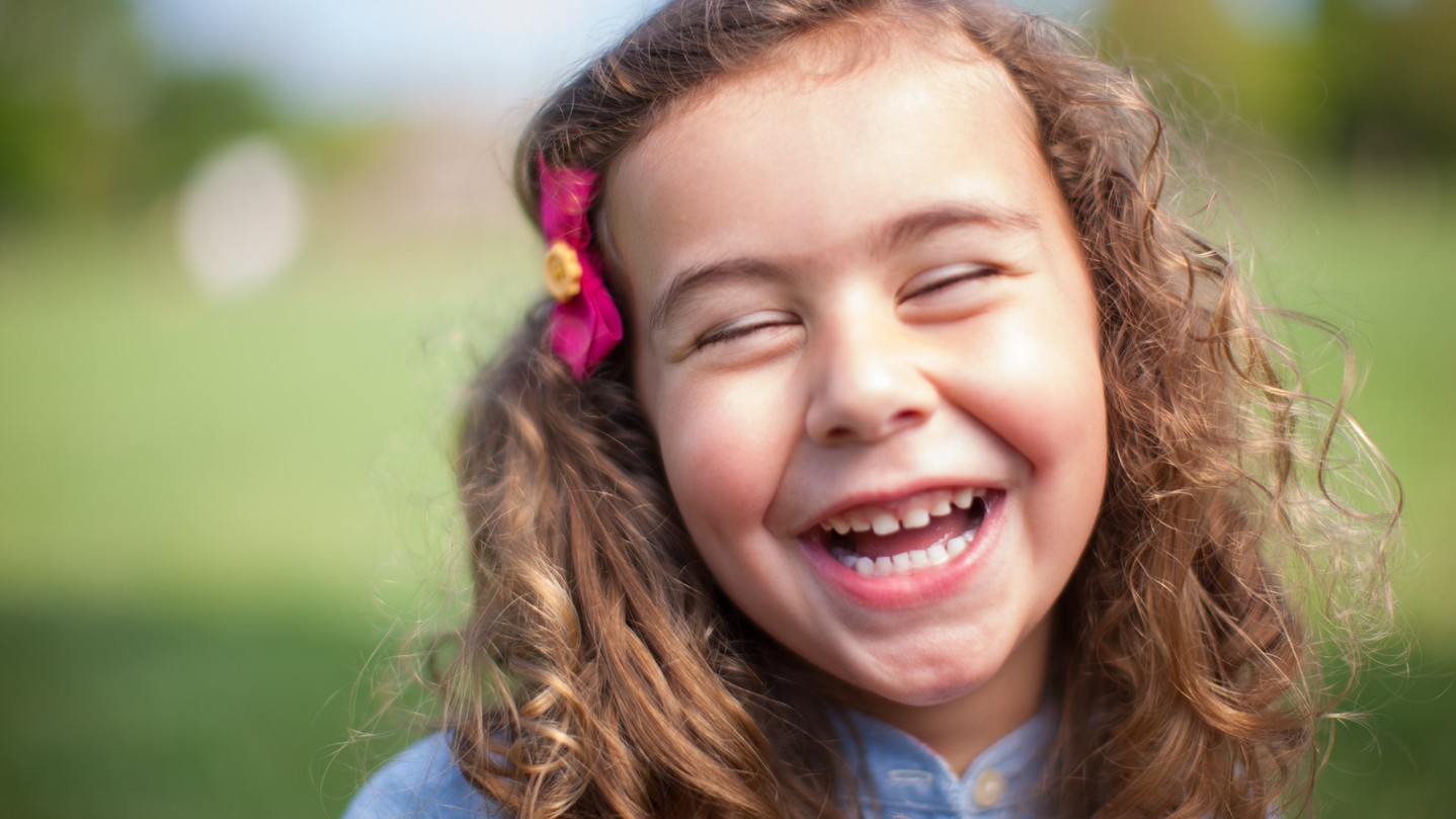 Close-up of a cheerful young girl with curly hair smiling widely outdoors in a grassy setting, wearing a pink bow.