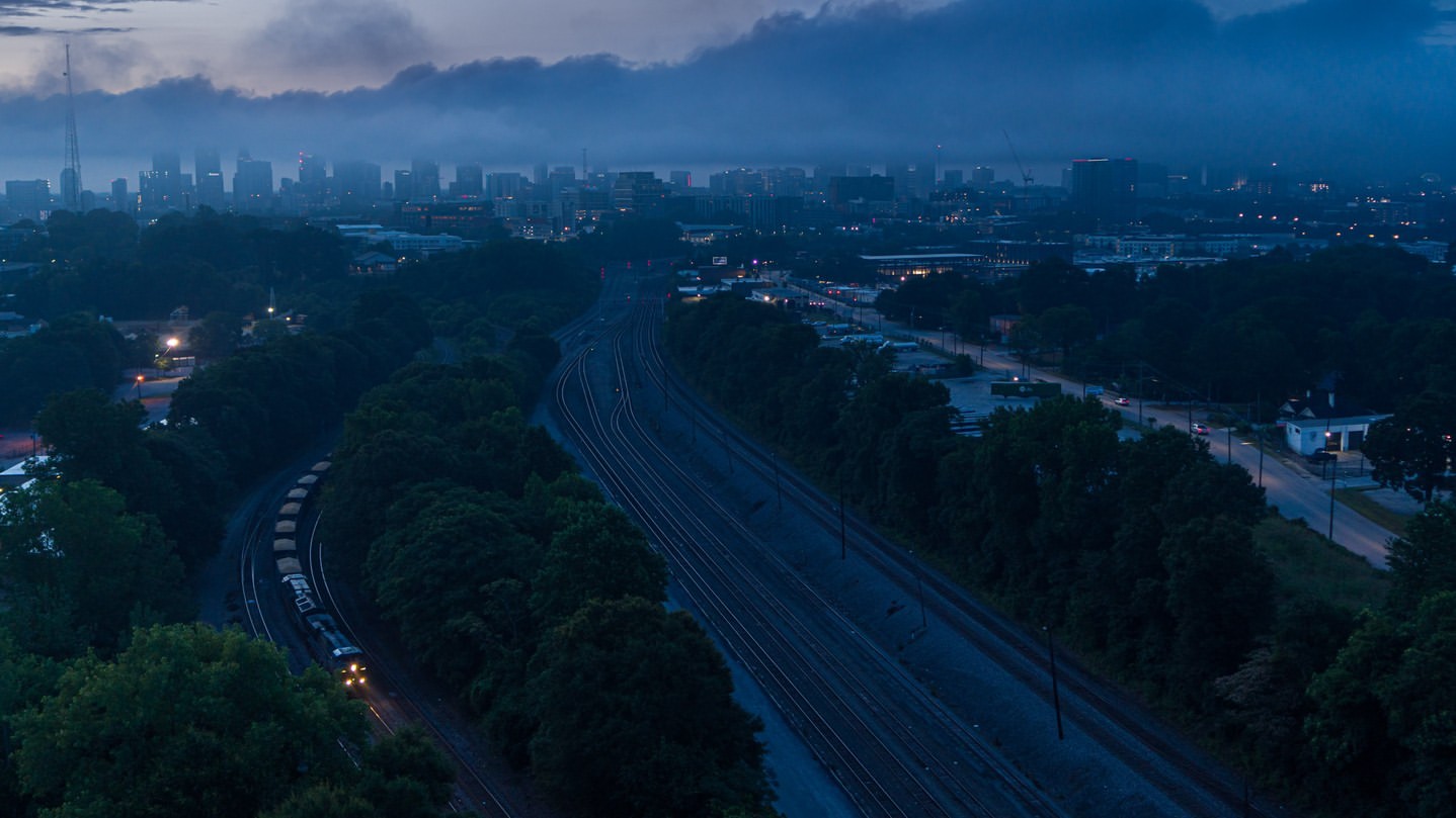 City skyline and railroad tracks at dusk. An aerial view reveals a train moving along the tracks with green trees surrounding the scene.