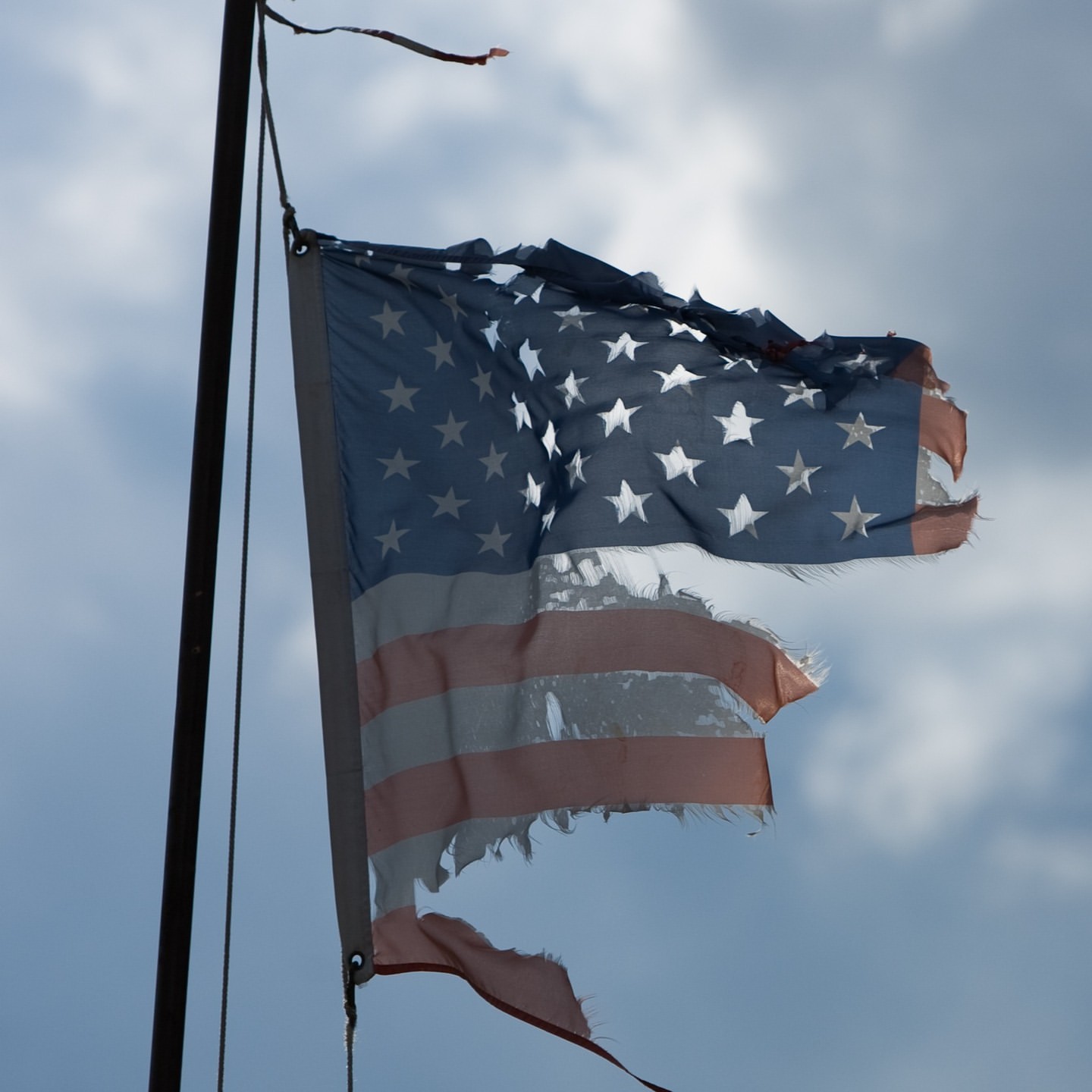 Tattered American flag waving on a pole against a cloudy sky, showcasing the flag's distressed condition and patriotic symbolism.