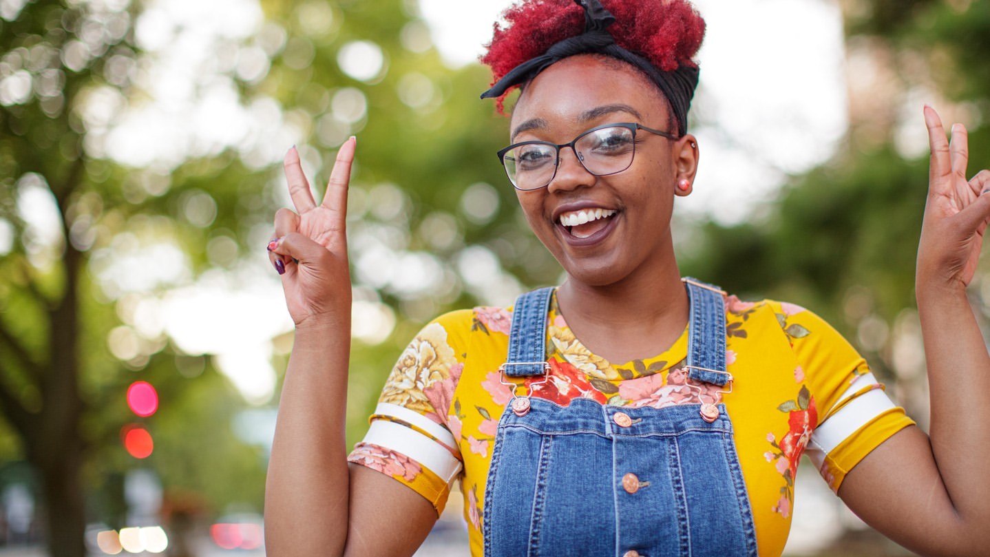Smiling woman with red hair and glasses making a peace sign outdoors against a green tree background. Portrait orientation, medium shot.