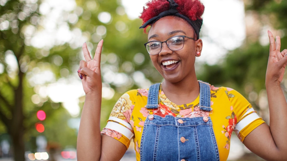 Smiling woman with red hair and glasses making a peace sign outdoors against a green tree background. Portrait orientation, medium shot.