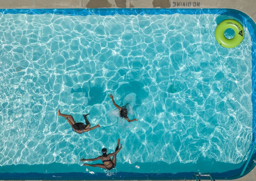 Aerial view of three women swimming in a clear turquoise pool with sunny reflections and a floating green inflatable ring. 