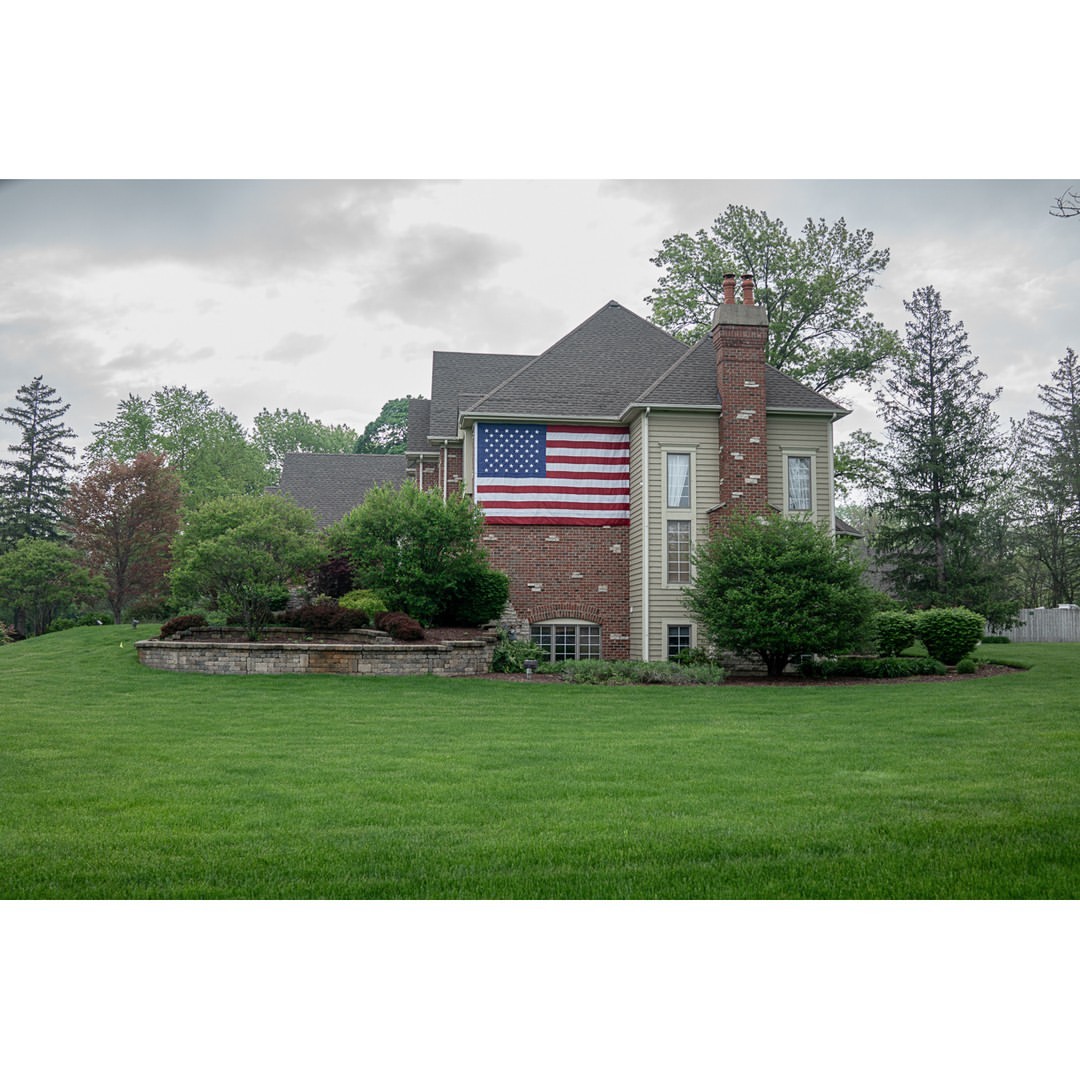 American flag displayed on the side of a large house with a green lawn and surrounding trees under an overcast sky.