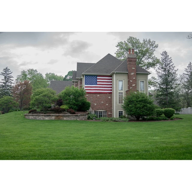 American flag displayed on the side of a large house with a green lawn and surrounding trees under an overcast sky.