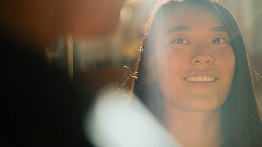 Close-up portrait of a smiling Asian woman with long dark hair in bright sunlight, shallow depth of field, and a blurry figure in the foreground.