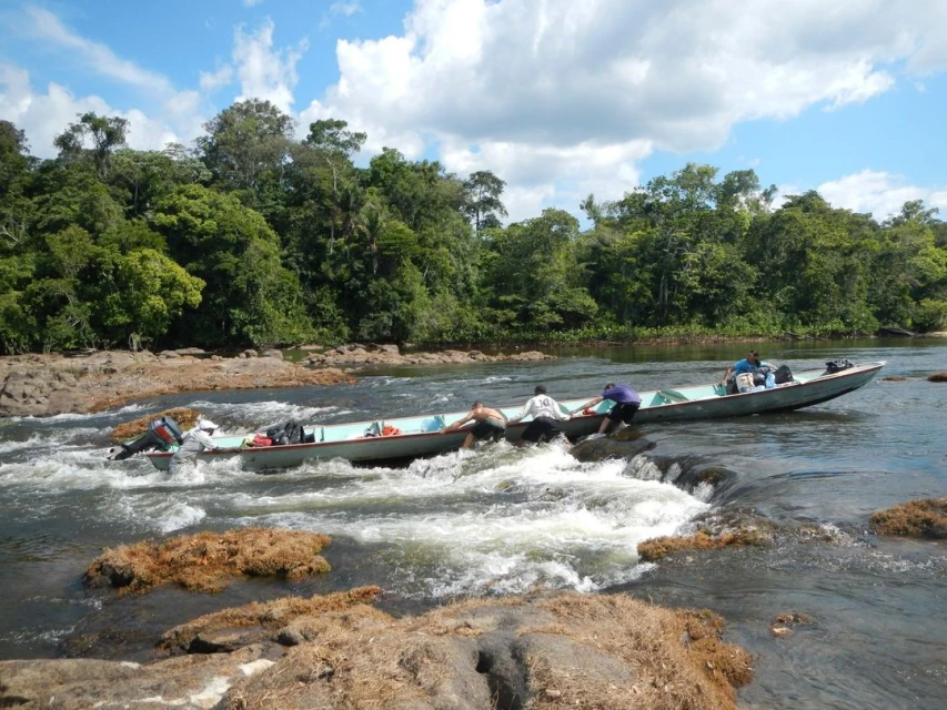 People maneuver longboats through the rapids on a rocky river surrounded by a lush green tropical forest under a partly cloudy sky.