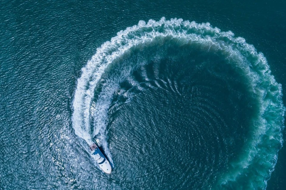 Aerial view of a white yacht sailing on dark blue water, creating a circular white wake pattern in the ocean.