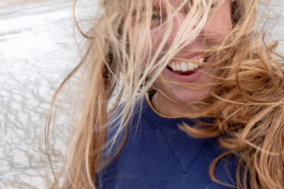 Close-up of a smiling blonde woman with hair blowing in the wind, wearing a blue sweatshirt against a blurred, neutral-toned background.