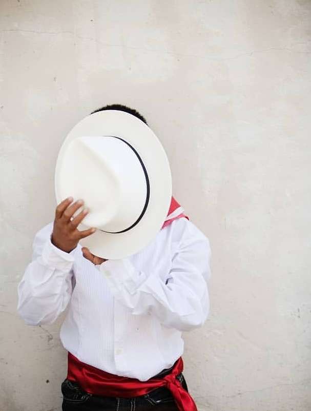 Young boy in traditional clothing hides his face behind a white sombrero against a neutral wall, showcasing cultural attire.