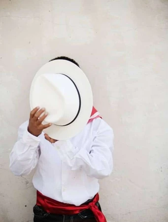 Young boy in traditional clothing hides his face behind a white sombrero against a neutral wall, showcasing cultural attire.