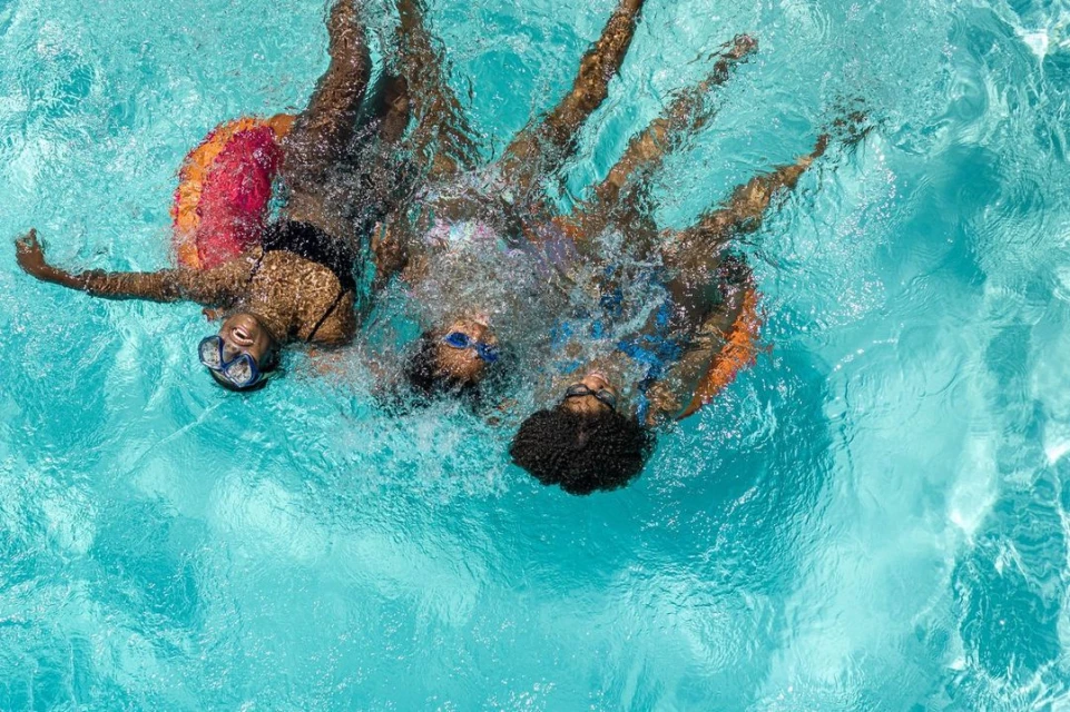 Overhead view of three young women of African descent swimming in a bright turquoise pool with swim rings and swim goggles.