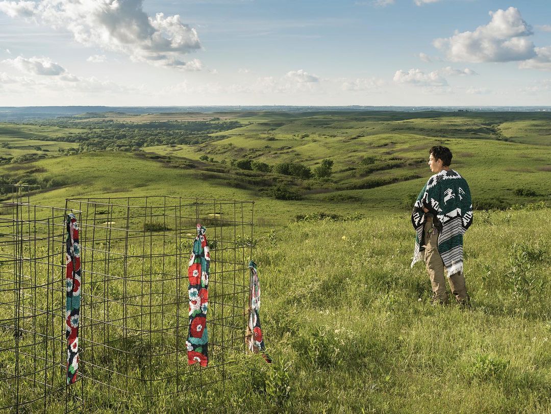 Man in poncho stands on grassy hillside overlooking a green prairie landscape with metal fencing and fabric strips in foreground on a sunny day.
