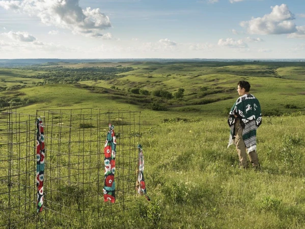 Man in poncho stands on grassy hillside overlooking a green prairie landscape with metal fencing and fabric strips in foreground on a sunny day.