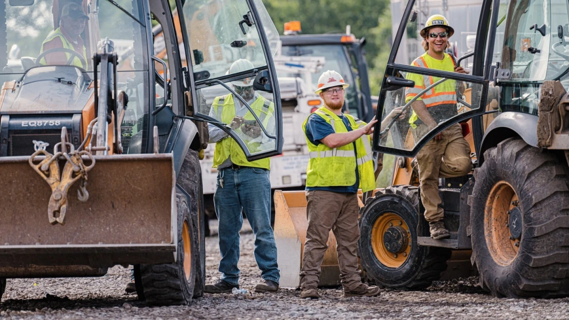 Construction workers stand near heavy equipment on a construction site, wearing hard hats and safety vests, working during the day.