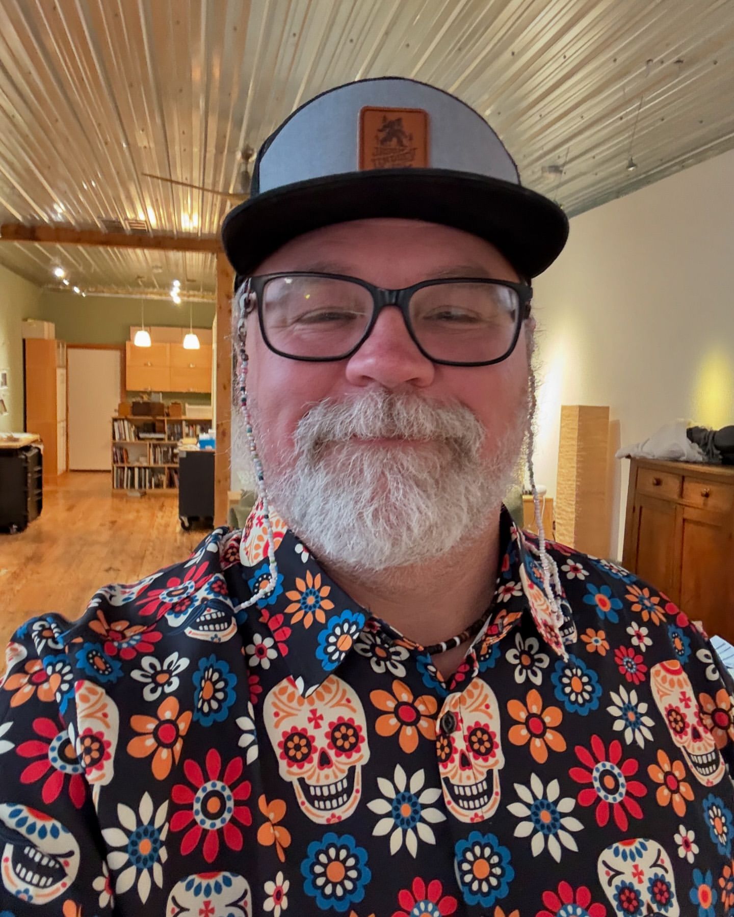 Adult male in a floral and skull shirt and ball cap stands indoors, wearing glasses and a short beard, with an indoor setting.