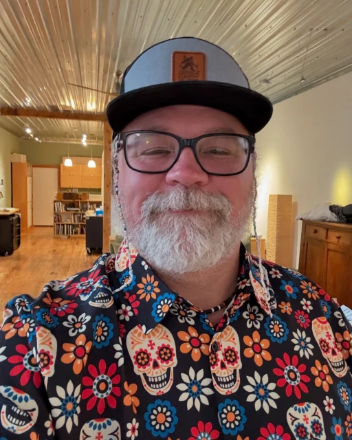 Adult male in a floral and skull shirt and ball cap stands indoors, wearing glasses and a short beard, with an indoor setting.