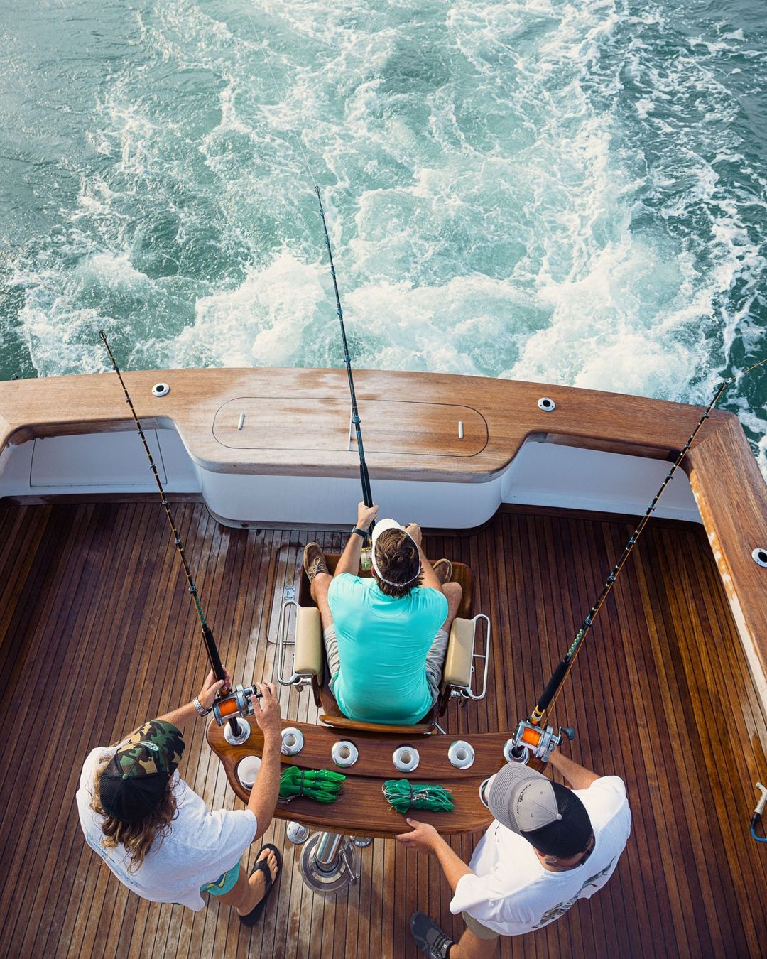 Three people fishing on a boat from an overhead angle, featuring fishing rods, wooden deck, and ocean water with wake.