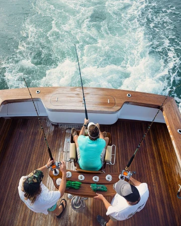 Three people fishing on a boat from an overhead angle, featuring fishing rods, wooden deck, and ocean water with wake.